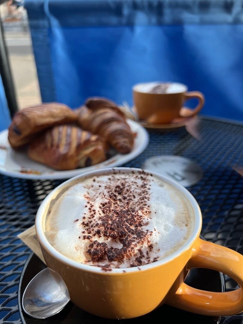 A cappuccino in a yellow cup dusted with chocolate, with croissants and a pain au chocolat on a plate behind, on an outdoor café table
